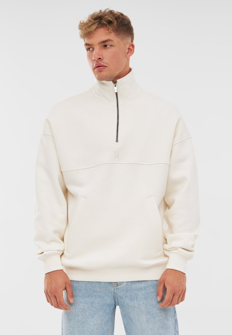 Light cream half-zip sweatshirt made from soft fabric, featuring a front pocket, ribbed cuffs, and a subtle logo on the chest.