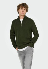 Dunkelgrüner Reißverschluss-Pullover mit Fischgrätmuster, lange Ärmel und geripptem Saum, getragen über einem weißen Hemd und hellgrauen Jeans.