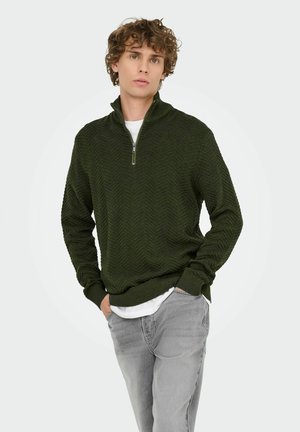 Dunkelgrüner Reißverschluss-Pullover mit Fischgrätmuster, lange Ärmel und geripptem Saum, getragen über einem weißen Hemd und hellgrauen Jeans.