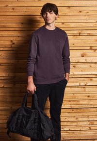 Bolsa de deporte negra con tela texturizada, correas ajustables y un bolsillo con cremallera, sostenida por una persona que lleva una sudadera morada oscura y pantalones negros.