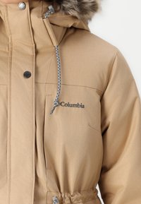 Svetlá zimná bunda s kapucňou s kožušinovým lemovaním, predným zapínaním na cvoky, sťahovacím pásom a logom "Columbia" na hrudi. Mäkká textúra.