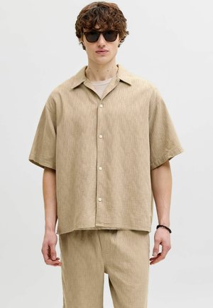 Young man with curly hair wearing black sunglasses, loose beige short-sleeve button-up shirt, matching pants, and a black beaded bracelet.