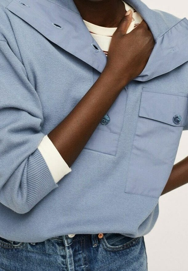 Persona che indossa una camicia azzurro chiaro sopra una maglietta bianca a maniche lunghe, con jeans blu, mano posata vicino al colletto della camicia.