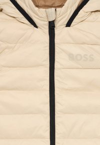 Beige boblejakke med glidelås foran og hette. Har quiltet struktur, svart glidelås og subtil "BOSS" logo på brystet.