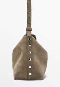 Taupefarbene Wildlederhandtasche mit einer vertikalen Reihe silberner Nieten und einem verstellbaren Riemen, präsentiert vor einem weißen Hintergrund.