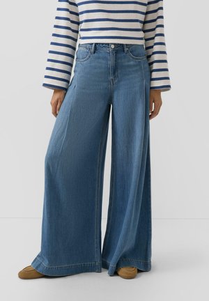 Personne portant un jean bleu à jambes larges, des chaussures marron et un haut blanc à manches longues avec des rayures horizontales bleu marine.