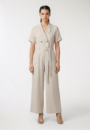 Donna con capelli intrecciati che indossa una tuta beige con maniche corte e cintura, e tacchi oro con punta aperta, in piedi davanti a uno sfondo bianco neutro.