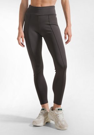 Kvinde iført sorte højtaljede leggings og hvide atletiske sneakers står mod en ensfarvet baggrund.