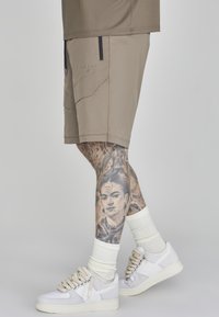 Beige Sportshorts mit Reißverschlusstaschen und Logodetails, kombiniert mit weißen Turnschuhen und hohen weißen Socken. Auffällige Tattoo-Kunst auf dem Bein sichtbar.