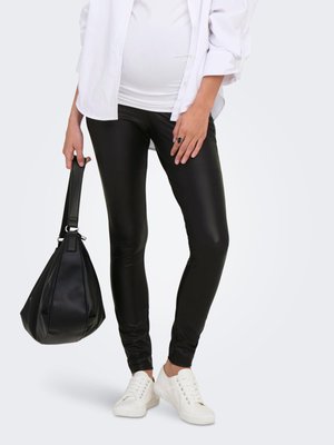 Persona embarazada vistiendo camiseta blanca, pantalones ajustados negros y zapatillas blancas, sosteniendo un bolso negro al lado.