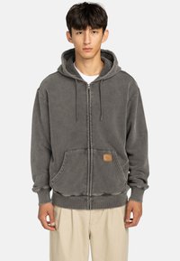 Sudadera gris con cremallera hecha de una tela suave con acabado texturizado, cuenta con un bolsillo frontal y un parche de cuero con la marca. Cuerdas unidas a la capucha.