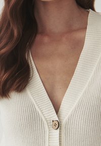 Cárdigan blanco acanalado con un profundo escote en V, que presenta un botón de madera en el frente, exhibiendo un diseño de punto texturizado.