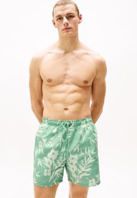 Grüne Blumenshorts mit elastischem Bund und Kordelzug. Sie weisen hellfarbige Blumenmuster auf, die sich gegen den einfarbigen grünen Hintergrund abheben.