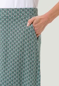 Main avec des ongles manucurés reposant à l'intérieur de la poche d'une jupe à motifs géométriques teal et blanche, portée avec un haut blanc.