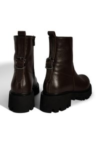 Bottines en cuir marron avec une finition texturée, présentant des zippers sur les côtés et des accents de boucles à l'arrière, reposant sur des semelles noires épaisses.