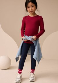 Haut rouge à manches longues, leggings navy, veste en denim nouée à la taille, baskets blanches, chaussettes rayées rouges et blanches, arrière-plan doux.