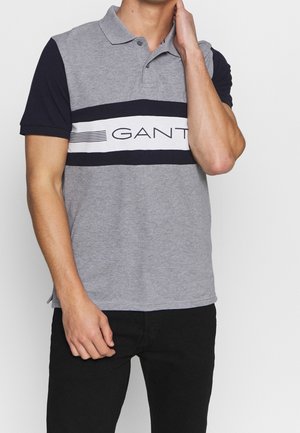 Man wearing a grey and black GANT polo shirt with short sleeves, standing with one hand touching his neck and black pants.