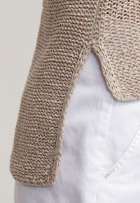 Gros plan d'un vêtement beige en maille, fente latérale sur un pantalon blanc avec une couture de poche visible et une petite étiquette en tissu.