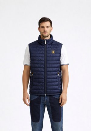 Uomo che indossa un gilet trapuntato navy sopra una camicia bianca a maniche corte, abbinato a jeans bicolore blu, in piedi davanti a uno sfondo bianco semplice.