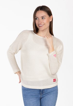 Mujer sonriendo, vestida con un suéter blanco de punto con un parche rojo que dice "LOVE" y jeans azules, posando frente a un fondo blanco liso.