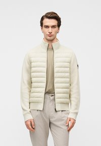 Ελαφριά μπεζ μπουφάν με σχεδιασμό εφαρμοστό, διαθέτει φερμουάρ μπροστά, ψηλό γιακά και μανίκια από υφασμένο ύφασμα με υφή. Φοριέται πάνω από μπεζ τοπ.