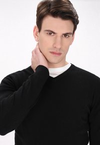 Maglione nero a collo a V realizzato in tessuto morbido, con maniche lunghe. Il modello ha un'espressione neutra e si sta toccando il collo.