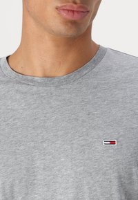 Tricou gri din bumbac cu decolteu la baza gâtului și un mic logo brodat în roșu, alb și albastru pe partea stângă a pieptului.