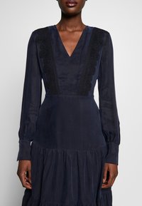 Marinblå klänning med V-halsringning och långa ärmar, har spetsdetaljer längs halsringningen och en flikig, volangkant. Slät textur.