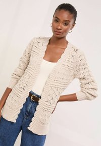 Vajszínű, horgolt kardigán, nyitott mintákkal, bordás mandzsettával és szegéllyel. Texturált dizájnnal kiemelve, fehér felsőre és kék farmerekre viselve.