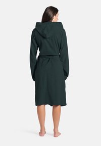 Robe con cappuccio di colore verde scuro, realizzata in morbido tessuto in spugna. Presenta una cintura in vita e maniche lunghe che arrivano a metà polpaccio.
