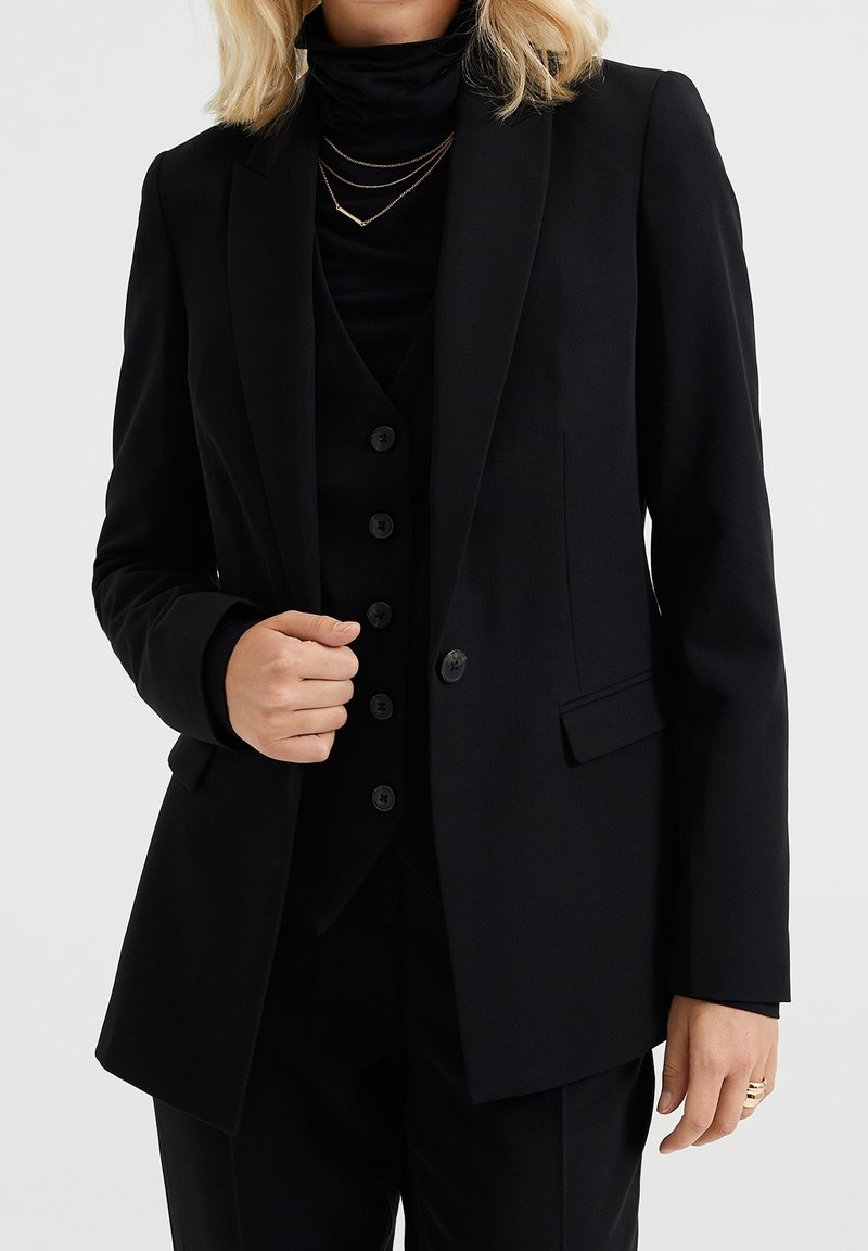 Svart skräddarsydd blazer med en mjuk textur, med en enkel knappstängning och två framfickor. Bärs över en svart polotröja.