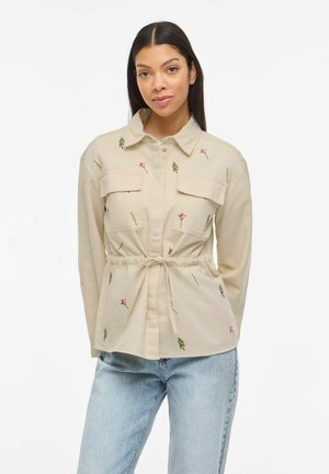 Femme portant une veste beige boutonnée avec broderies florales et ceinture nouée, associée à un jean bleu clair, debout devant un fond blanc.