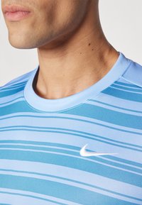 Lys blå Nike treningsskjorte med horisontale striper i varierende nyanser av blått. Har rund halslinning og hvit swoosh-logo.
