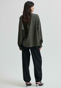 Camicia verde oliva oversize con maniche lunghe, polsini a bottone, orlo arrotondato, abbinata a pantaloni neri larghi e scarpe con tacco nere.