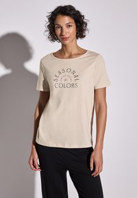 T-shirt de color beige claro con mangas cortas, escote redondo y texto negro que dice "COLORES DE TEMPORADA". Material de algodón suave con un corte relajado.
