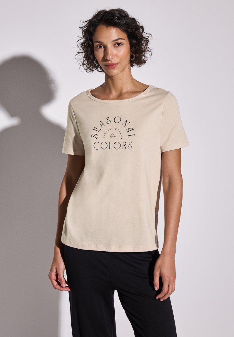 T-shirt de color beige claro con mangas cortas, escote redondo y texto negro que dice "COLORES DE TEMPORADA". Material de algodón suave con un corte relajado.