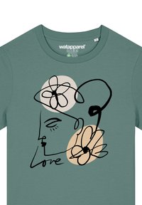 T-shirt en coton vert avec un dessin au trait noir d'un visage entouré de fleurs et du mot "amour", accentué par des cercles beiges.