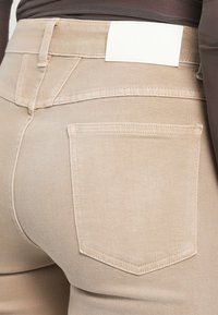 Gros plan d’un pantalon beige montrant une poche arrière, une passants de ceinture et une étiquette blanche, porté avec un haut marron foncé.