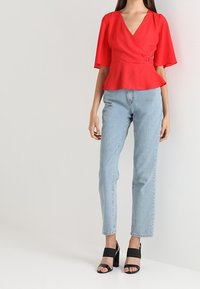 Kvinde iført en rød kortærmet omslagstop med bælte detalje, lyseblå højtaljede jeans og sorte åbne hæle med blokhæle, stående op ad en hvid væg.