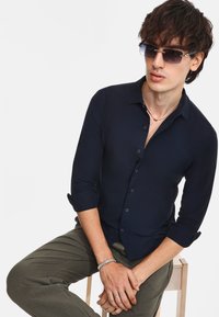 Camicetta aderente blu navy con colletto, maniche lunghe arrotolate, abbinata a pantaloni verde oliva. Modello che indossa occhiali da sole e accessori.