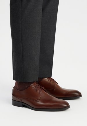 Zapatos de vestir de cuero marrón con un acabado suave, con cordones y un tacón ligero, combinados con pantalones de traje gris oscuro y calcetines con estampado.
