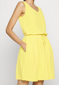 Gul ärmlös klänning med V-halsringning, elastisk midja, sidofickor och en svävande kjol. Tillverkad av mjukt, lätt material.