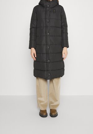 Personne portant un long manteau matelassé noir avec capuche, un pantalon beige à jambes larges et des chaussures marron, debout devant un fond clair uni.