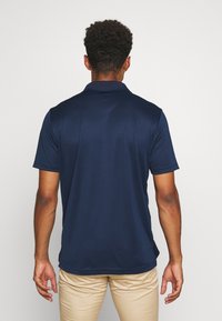 Osoba stojí zády kameře, má na sobě tmavě modrou polo košili a béžové kalhoty, na neutrálním pozadí.