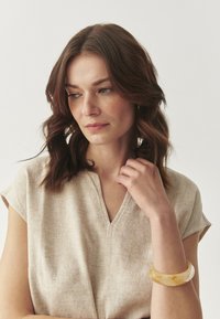 Top beige in lino con scollatura a V e maniche corte; indossa un bracciale traslucido color ambra sul polso.