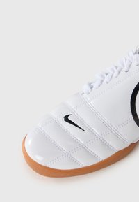 Chaussure de sport Nike blanche avec logo swoosh noir et semelle marron, présentant des motifs cousus et des lacets blancs, sur fond gris clair.
