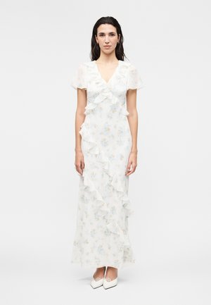 Γυναίκα στέκεται φορώντας λευκό φλοράλ μάξι φόρεμα με λεπτομέρειες βολάν και φαρδιά μανίκια, συνδυασμένο με λευκές μυτερές γόβες.