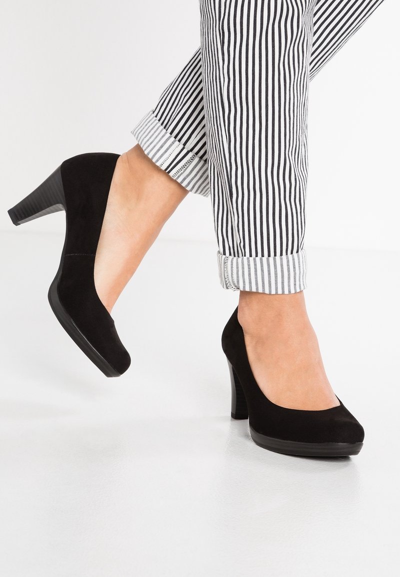 Femme portant des chaussures à talons carrés en suède noir, coupées à la cheville, avec un pantalon rayé noir et blanc retroussé aux ourlets.