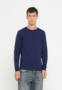 Tamsiai mėlynas ilgos rankovės T-shirt, pagamintas iš medvilnės, su apvaliu iškirpimu ir prigludusiomis rankovėmis, dėvimas su šviesiai mėlynomis džinsais.