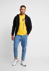 Sort hættetrøje med lynlås over en gul t-shirt med et palmetræ grafik, parret med lyseblå jeans og hvide sneakers.